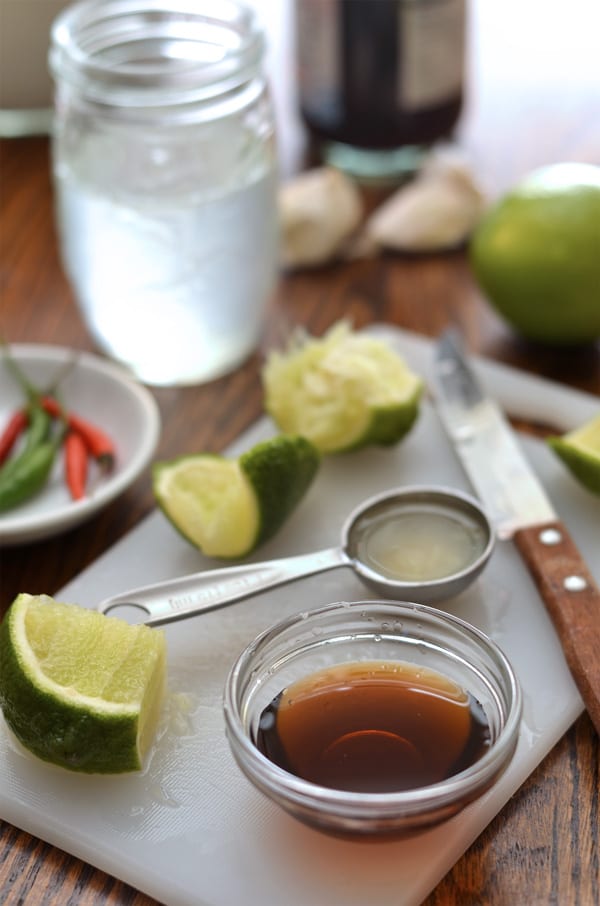 A small glass bowl of amber fish sauce beside lime wedges, chiles, garlic, water, and a measuring spoon.